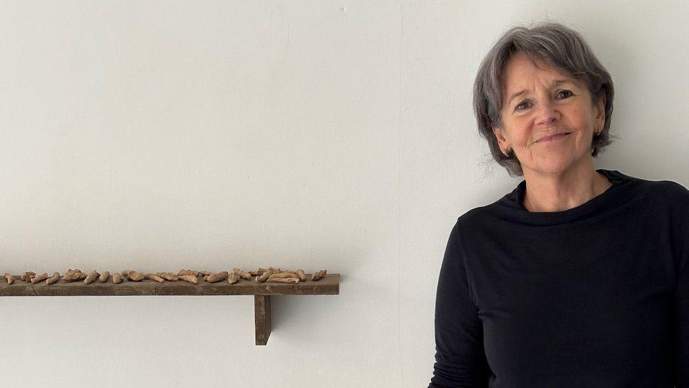 Helen stood to the right, against a white wall. She is smiling. There is a shelf to her left with pieces of her artwork displayed.
