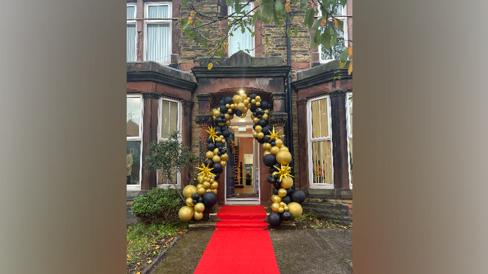 Black and gold balloons frame a Victorian doorway of a house, a red carpet is coming from the front door into the front garden, the front door is open