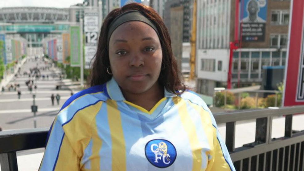 A woman wearing a blue and yellow football shirt stands beside a railing overlooking Wembley Way, with Wembley Stadium visible in the distance. Large “TOGETHER” banners line the walkway behind.