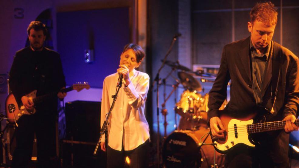 Cocteau Twins performing on stage. Elizabeth Fraser is singing into a mic and there are band members on guitars and drums 
