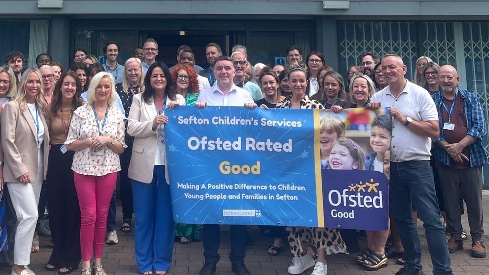 Members of staff from Sefton Council's children's services huddled together smiling and holding a banner which reads 'Ofsted rated: good'.