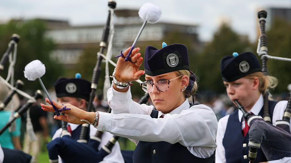 A woman in colourful glasses marches with a pipe band. She is twirling tenor drum sticks in the air