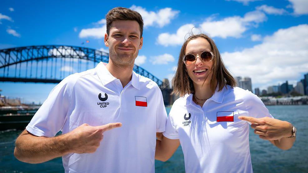 Hubert Hurkacz and Iga Swiatek point at each other while grinning and wearing white Poland team polo shirts, with Sydney Harbour Bridge in the background
