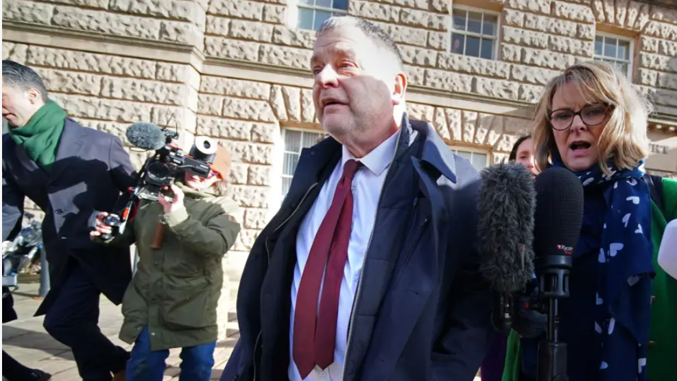 Mike Amesbury, wearing a navy blue coat over a white shirt and burgundy tie, walks away from court flanked by camera operators and journalists.