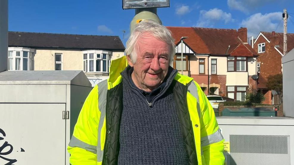 An man with white hair wearing a high visibility coat over a navy blue jumper. He is looking at the camera. Only the top half of his body is visible. You can see a row of houses behind him and a blue sky.