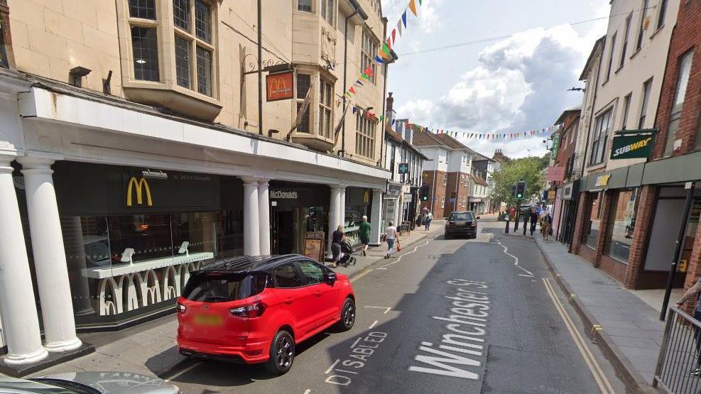 A view of Winchester Street on a sunny day with a red car parked in a disabled bay outside of the McDonald's on the left