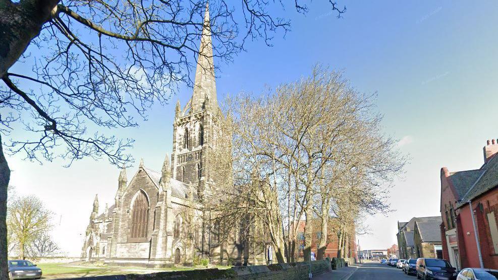 A large church with white and grey brick. The sky is blue and the building is surrounded by trees and green space. A road is on the right hand side. 