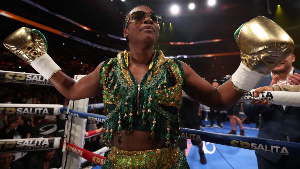 Claressa Shields holds out her arms and is wearing gold boxing gloves and a gold and green vest as she stands next to the ropes of a boxing ring
