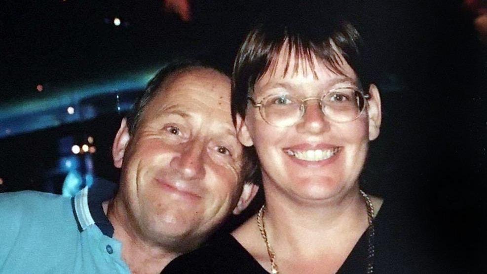 A man and a woman smile at the camera. The woman is wearing glasses