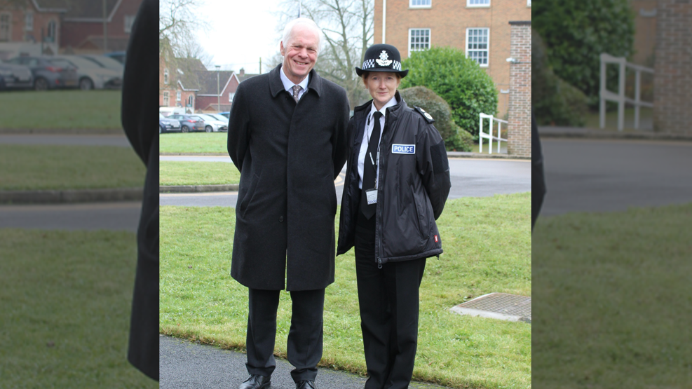 two people standing next to each other looking at the camera in front of a grassy verge with buildings in the background. The woman on the right wears a police hat, coat and tie, while the man on the left wears a heavy overcoat