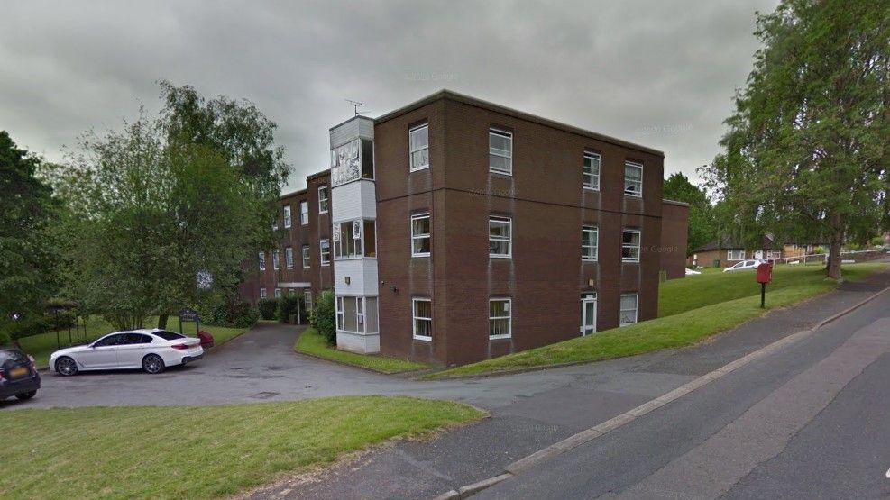 A two story brown brick building with shite windows. It is next to a road and sits next to green grass, with cars parked to its left