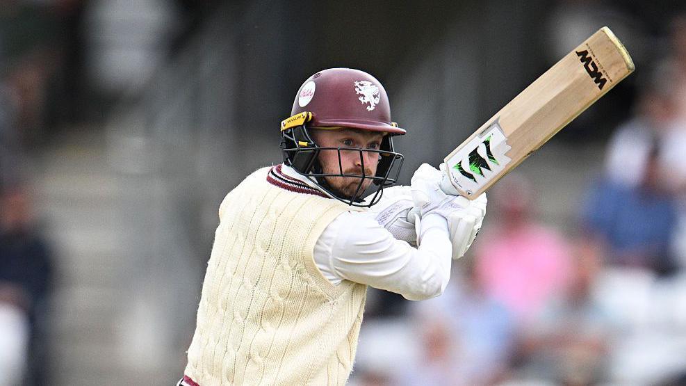 Lewis Goldsworthy in white cable jumper and maroon batting helmet holds his bat up to his right as he plays a shot