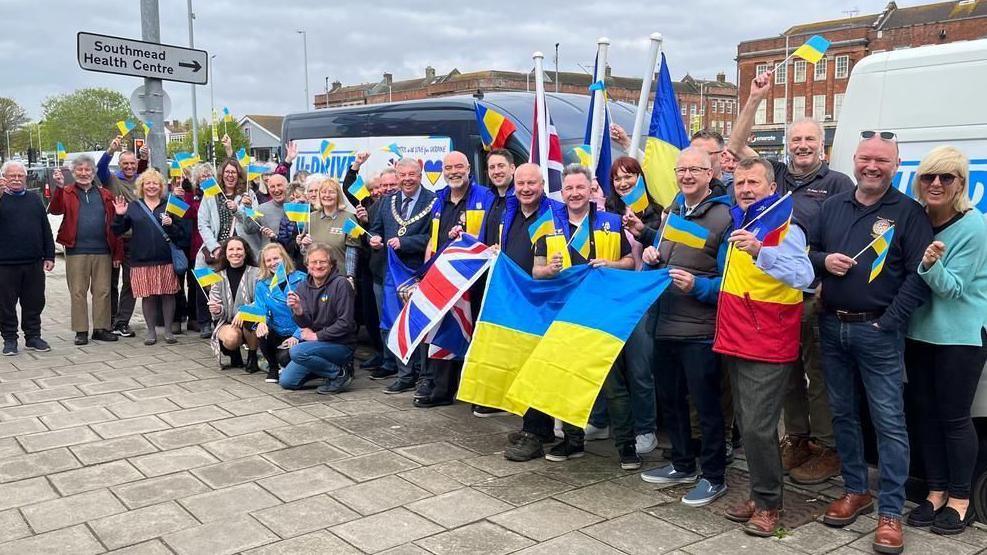 A crowd of people standing in front of two white vans. They are carrying British and Ukrainian flags and are all smiling at the camera