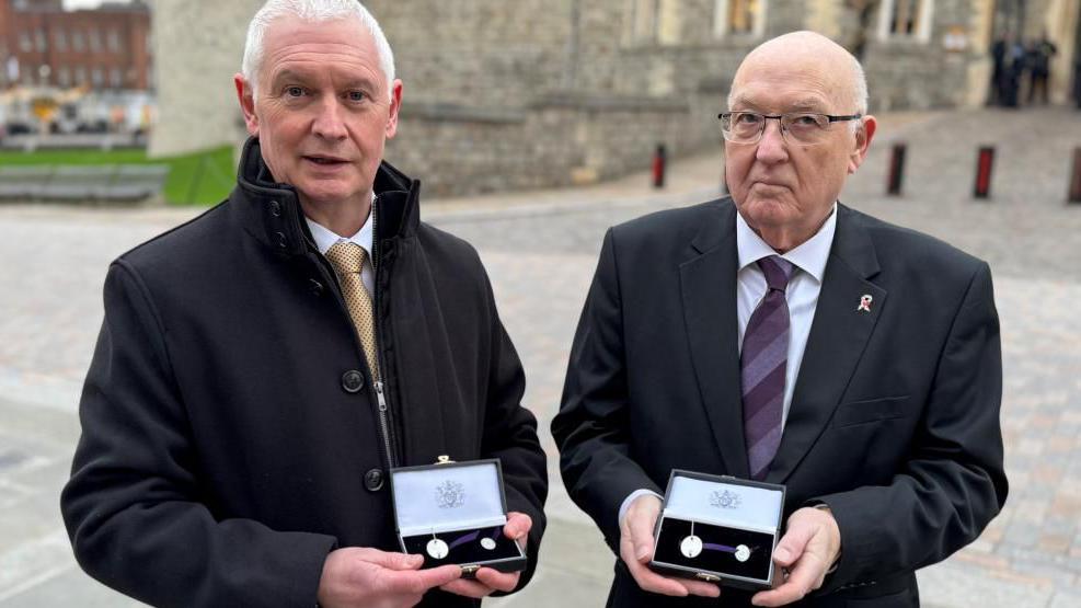 Two men wearing suits hold little boxes which contain silver emblems. 