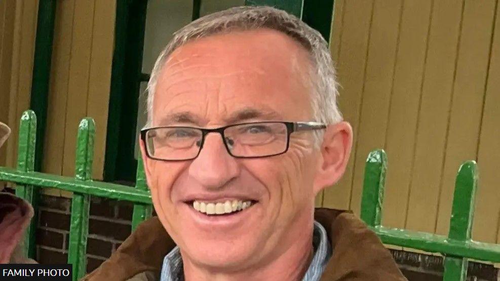 Islwyn Owen has short grey hair and is wearing glasses and a brown coat. He is smiling at the camera. Green painted railings and a brick and wood panelled wall can be seen behind him.