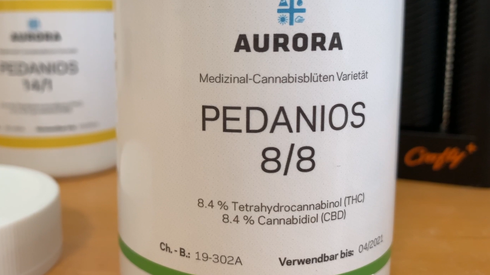 A white medicine bottle with the word 'Aurora' at the top and 'Medizinal-Cannabisluten Varietat. At the centre it has the words 'Pedanios 8/8' on it.