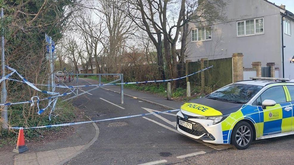 A police car is on the right of the picture, sitting in front of a road that has blue and white police tape across it. The small road has trees either side, and houses are behind the fences.