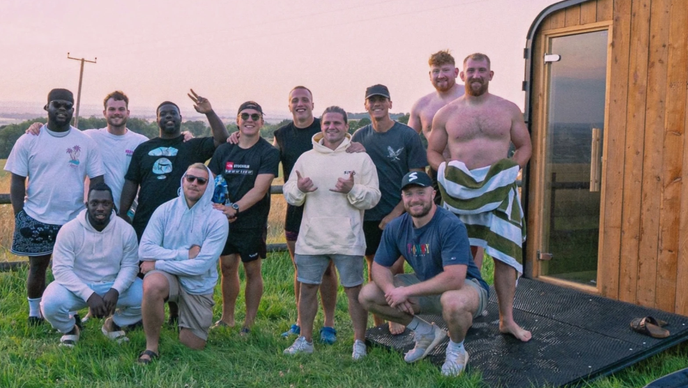 A group of men, some semi-clothed holding towels, pose outside a mobile sauna. They stand on a grassy field and there is a cold water plunge bath nearby. 