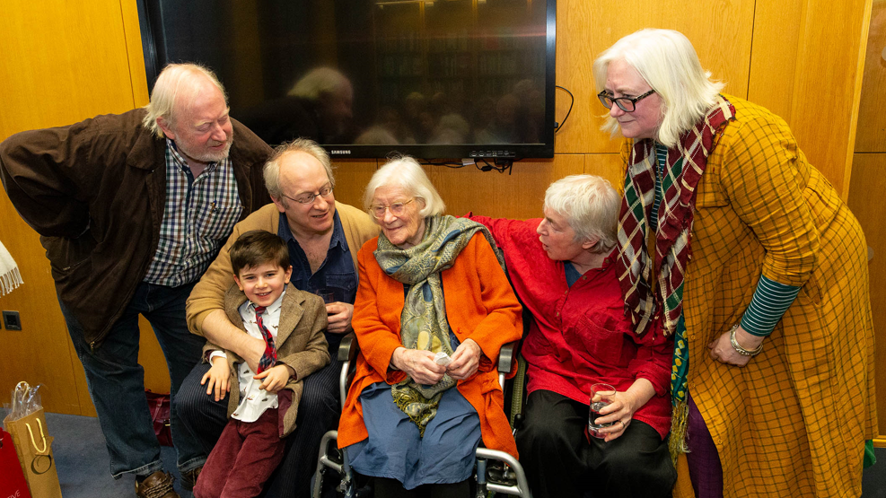 Shows Jennifer Johnston in a bright orange top with a green scarf sitting in a wheelchair with two men and a boy to the left and to women - one in a red top and the other in an orange top to the right. 