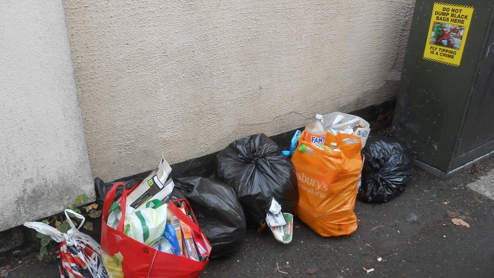 Several tied bags of rubbish are pictured on the floor in a street. A poster warning about fly-tipping is stuck on a green box 
