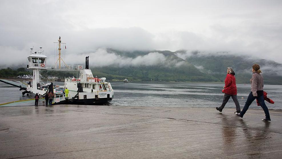 The Corran Ferry is a white and back boat. It is berthed at a slipway. Two women walk down the slipway towards the ferry. Mist hangs on the hills on the opposite shore.