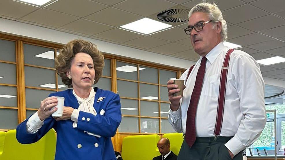 A woman dressed as Margaret Thatcher, wearing a blue jacket, on the left hand side, next to Nick, wearing a white shirt, red suspenders and a red tie