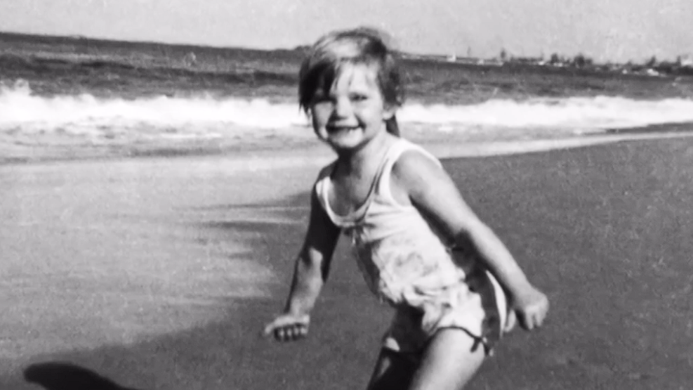 A black and white photograph showing three-year-old Cheryl Grimmer standing on a beach with waves gently lapping in the background. She has short fair hair and is wearing a white bathing costume. 