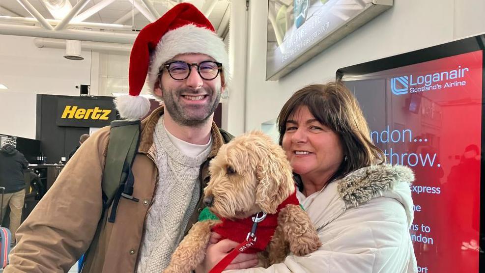 Deirdre has short brown hair. She has a beige coat on. She is holding her dog Skye is blonde and wearing a red Christmas jumper. Odhran is wearing a Santa hat and has dark framed glasses and stubble. He his wearing a brown coat and beige woolly jumper. 