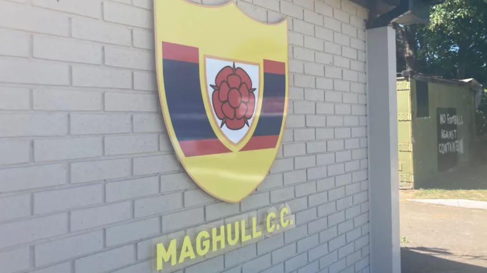 The yellow, black and red logo for Maghull Cricket Club set against a brick wall on a sunny day.