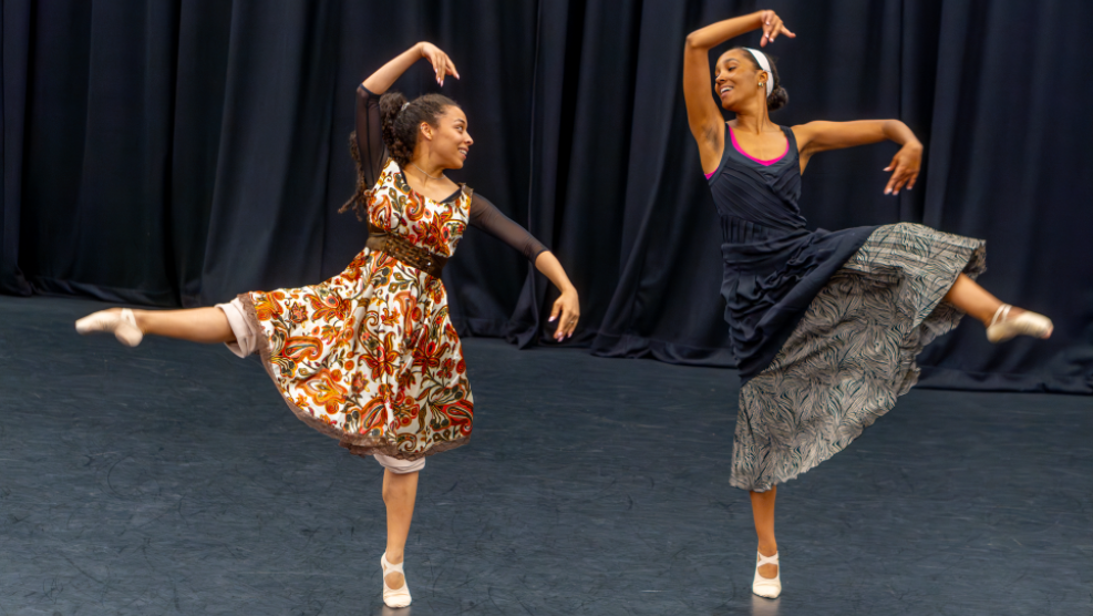 Two black women performing ballet on stage. They are both wearing long dresses and looking towards each other. They are posed forming shapes with their arms and kicking one leg out while standing on their toes.