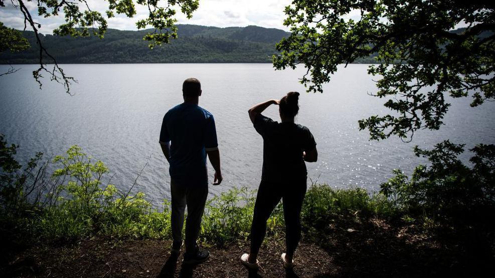 A man and woman stand on a leafy shoreline of Loch Ness. The woman shades her eyes with a hand as she and the man look out across the loch.