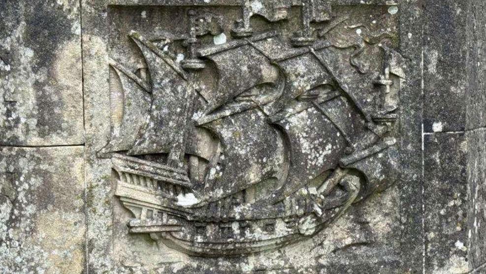 A detailed stone carving of a multi-masted sailing ship is embedded in a weathered stone wall. The surrounding stone is aged and mottled with lichen, suggesting long exposure to the elements.
