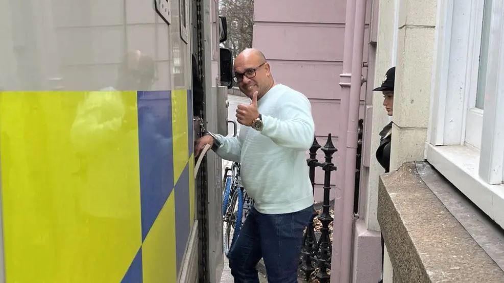 Gavin Neil Roberts pictured entering a police van. He is seen looking directly at the camera while holding his thumb up. He has a bald head and dark, rectangular glasses. He is mostly clean shaven and wearing a mint coloured jumper and blue jeans.