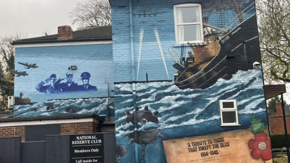The side of a large house-shaped building painted with a colourful mural. It depicts a black and brown trawler called Lifeguard in rough seas. Floating in the sea are round black mines with nodules and chains. Bombers fly in the sky and searchlights rise from land, to either side of a tall dock tower. Three men in naval uniform salute as they look out from the painting. A large red poppy is painted next to the name of the mural, "A Tribute to Those that Swept the Seas 1914 to 1945". A black sign can also be seen with the words National Reserve Club, members only, in white lettering.