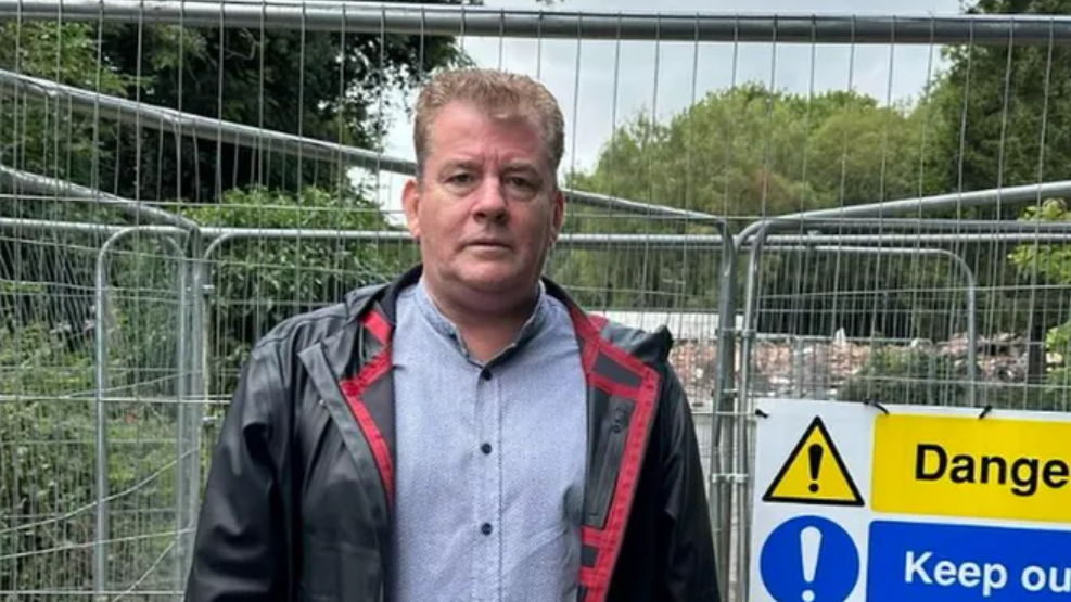 A man wearing a blue shirt and black and red jacket stands in front of the remains of the crooked house. There are gates and rubble behind him, as well as Danger and Keep Out signs.