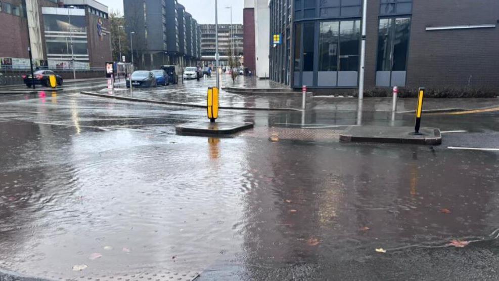 Water on the road surface at a junction with yellow bollards in the middle of the road and tall buildings behind