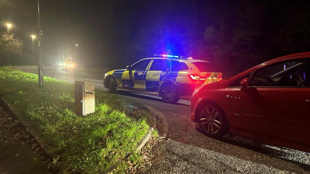 A police car with blue sirens flashing at the scene of the crash. A red car can also be seen in the photo.