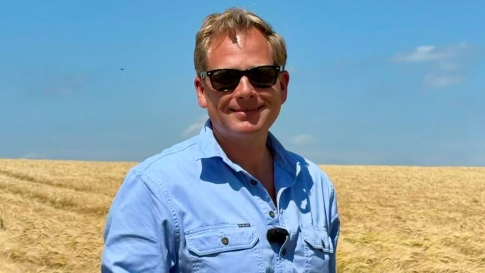 James Peck has short blond hair and is wearing black sunglasses and a blue shirt. He is standing in a field of yellow crops, and the sky is blue.