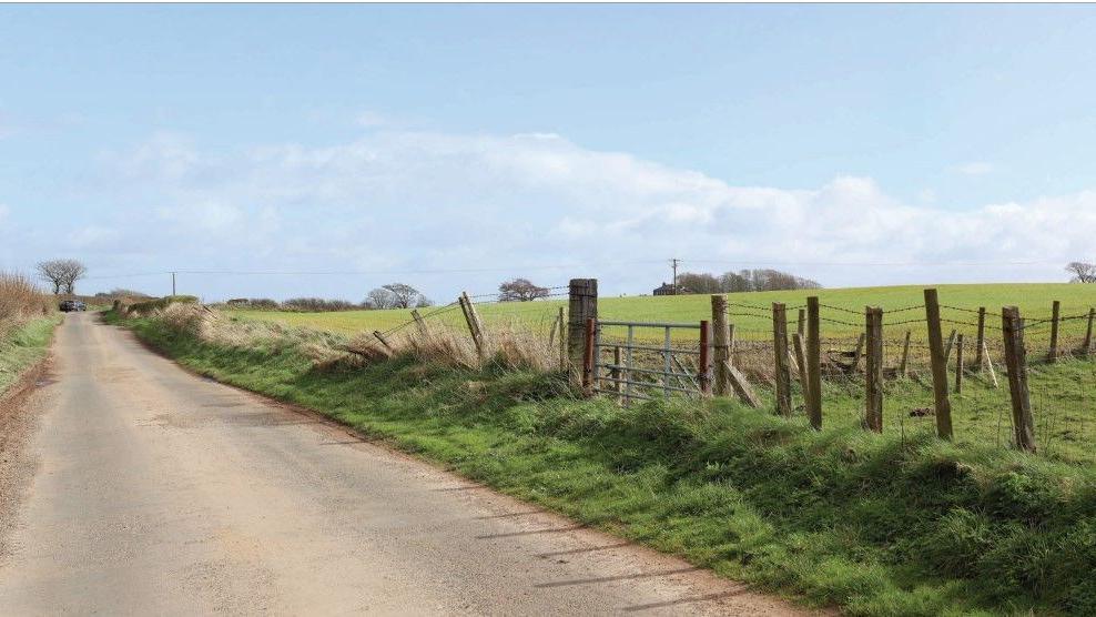 Image shows a rural road with a field to one site and a fence in need of repair