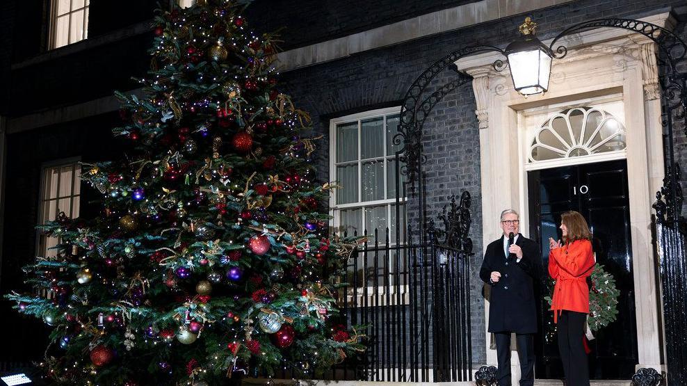 The grand Downing Street tree adorned with decorations, outside the front door of 10 Downing Street, where Sir Keir Starmer and his wife are stood