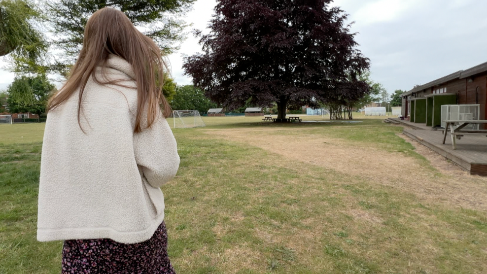 Ms Rhodes is wearing a white fluffy coat, she is walking across the school field with football goalposts and several large trees ahead of her, and low brick buildings to the right.