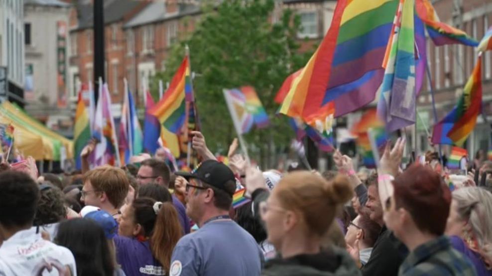 A crowd of people holding rainbow flags in a city street, standing in profile.
