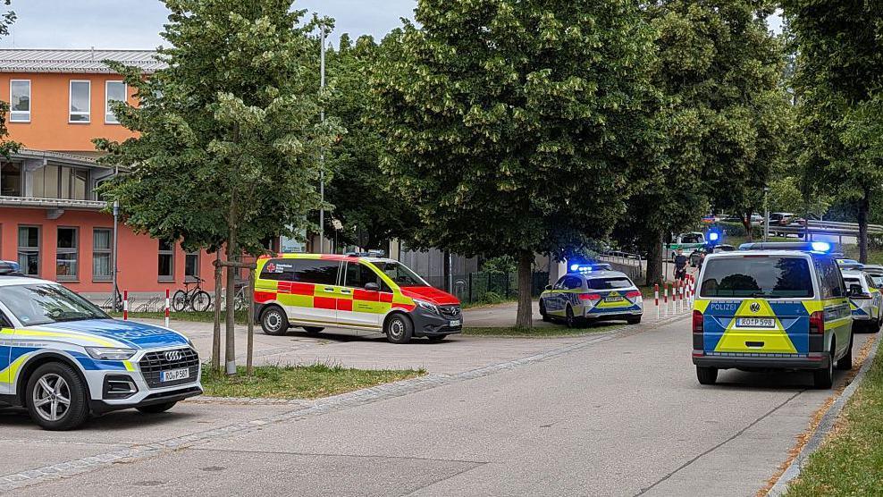 Emergency service vehicles with flashing lights outside a school building, the road is quiet.