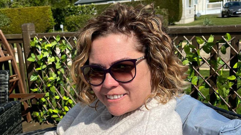A close-up image of a woman wearing sunglasses. She is smiling and looking into the camera.