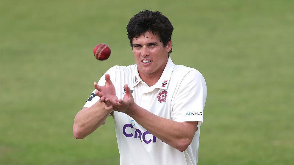 Liam Guthrie, wearing a white Northamptonshire shirt, cups his hands ready to catch the red cricket ball