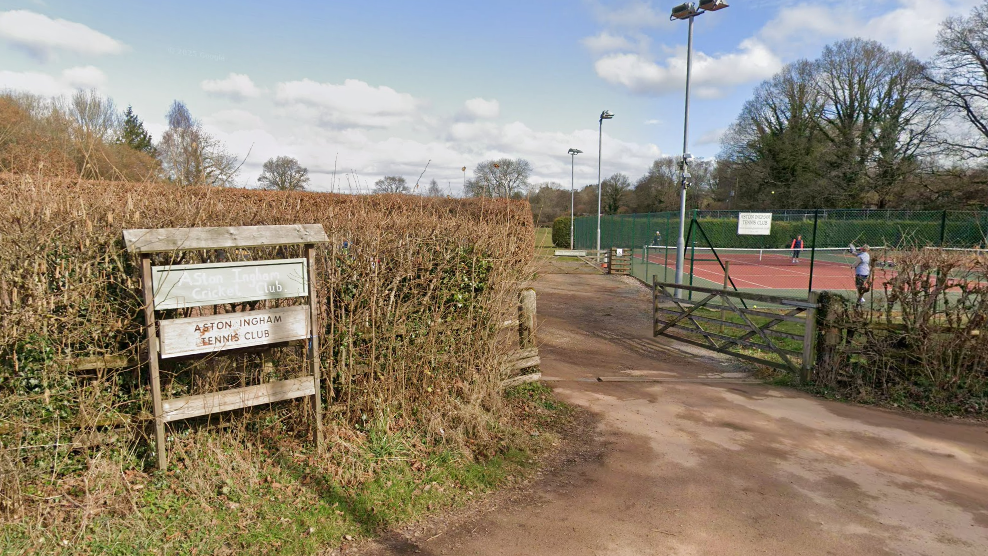 A wooden sign in front of a hedge saying Aston Ingham Cricket Club and Aston Ingham Tennis club. To the right of the picture behind a gate is a red tennis court with a few people standing on the court