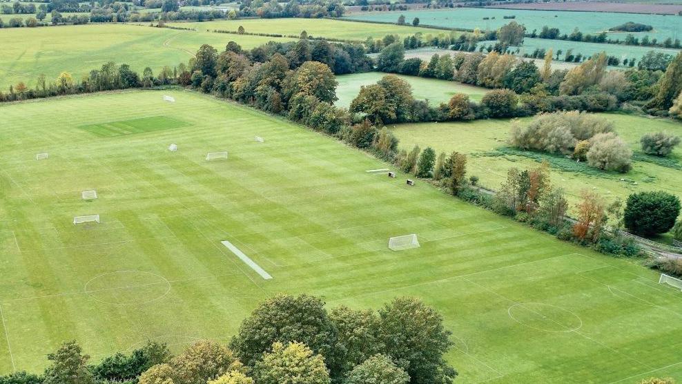 An aerial view of playing fields bordered by trees and hedging. The main field has football nets on it.