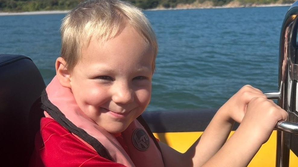 Benjamin with red polo shirt and pink life jacket on a sea motor launch. He has blonde hair.