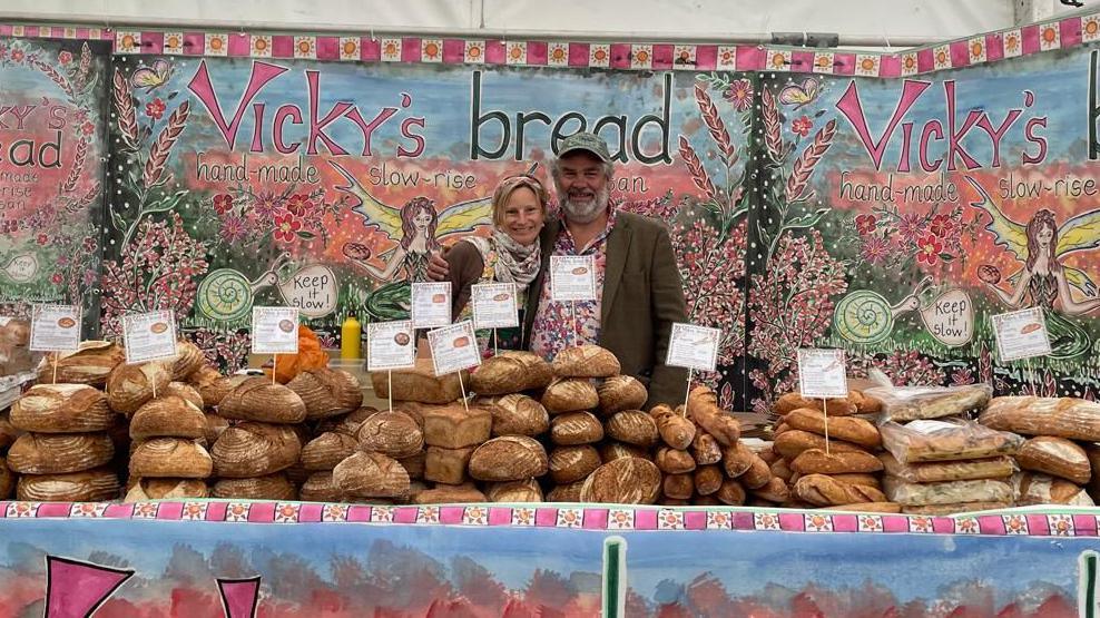 Vicky and Hairy Harford stand behind a long table loaded with loaves of bread