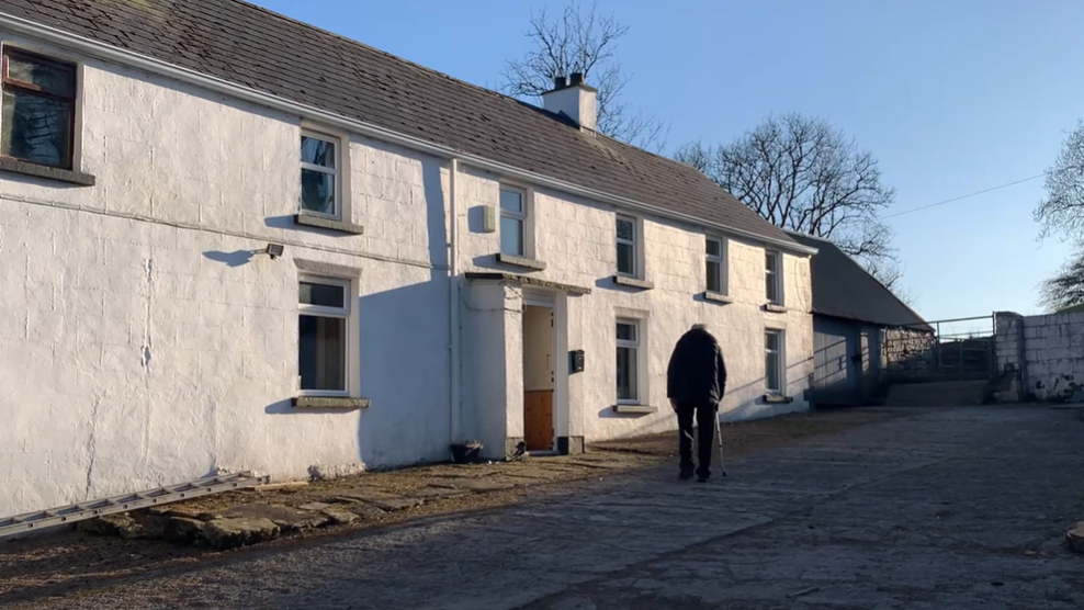 Owney walking towards the door of his house. He is wearing dark trousers and coat and has a walking stick. The farmhouse is white and there are sheds beside it with a gate.